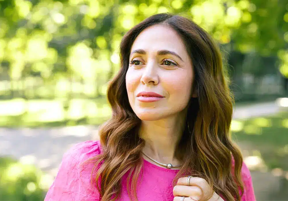 A hispanic woman wearing a pink shirt and staring into the foreground outside in a park.