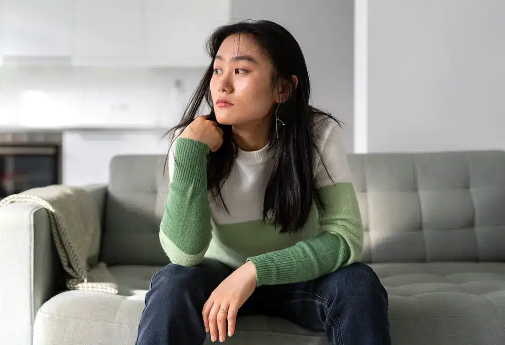 A depressed asian woman hunched over and looking outside while sitting on her couch.