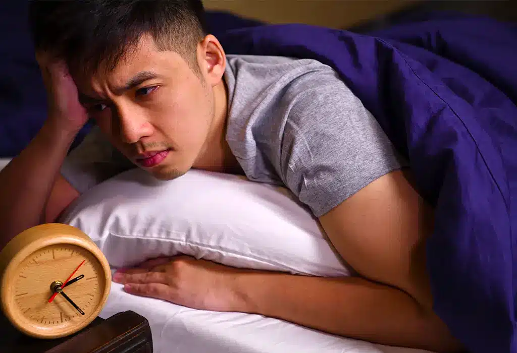 A distressed teenage boy staring at his alarm clock in bed.