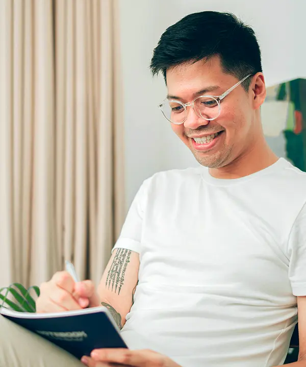 An asian man in his late 30s writes in his journal, smiling and focusing on his mental health.
